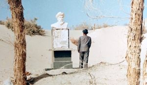 fotografia, colori, esterno, deserto peino sole, uomo di spalle con cappello osserva dal basso statua bianca di uomo con cappello