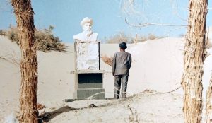 fotografia, colori, esterno, deserto peino sole, uomo di spalle con cappello osserva dal basso statua bianca di uomo con cappello