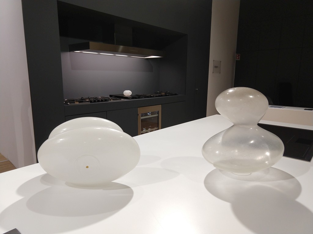 fotografia, colori, interno, due opere in vetro appoggiate su banco da cucina bianco, sfondo cucina e parete grigia, mostra "ab - L'Essenza dell'Assenza"