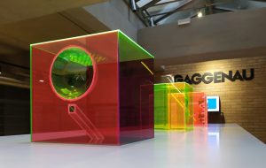 fotografia, colori, interno, 3 cubi di plexiglas colorato rosso, verde, giallo appoggiati su un grande tavolo bianco, sfondo mattoni e scritta gaggenau sul muro