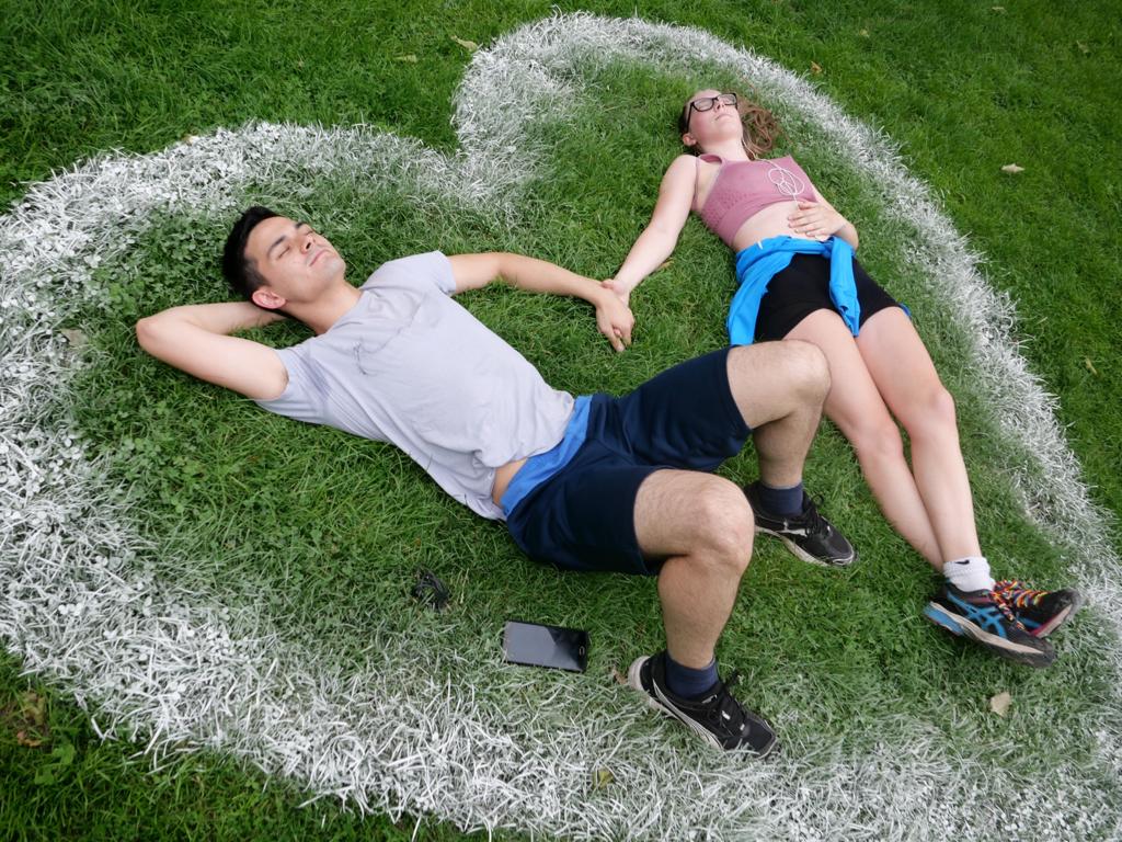 un ragazzo e una ragazza sdraiati sull'erba nel parco di Bristol all'interno del cuore bianco disegnato nell'erba che si tengono per mano