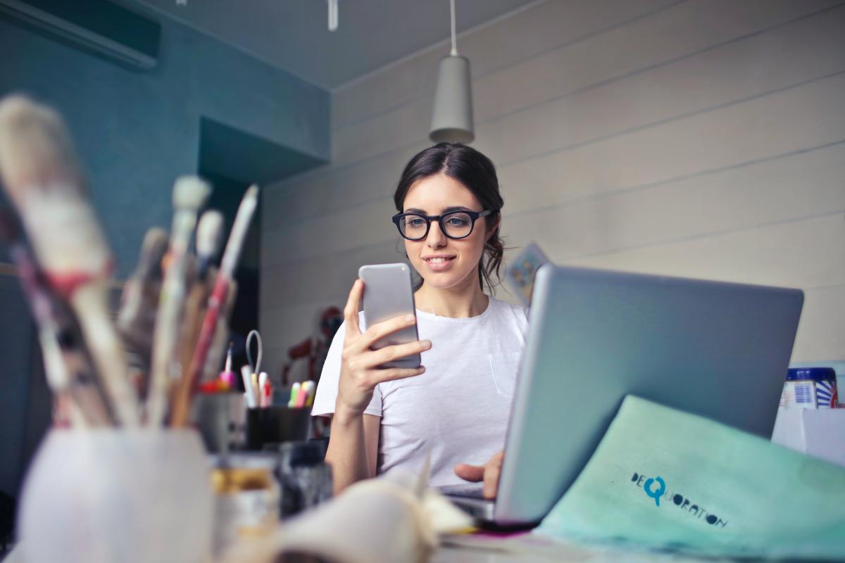 I migliori lavori da fare in smart working Ragazza con occhiali siede davanti ad un computer portatile mentre tiene nella mano destra uno smartphone