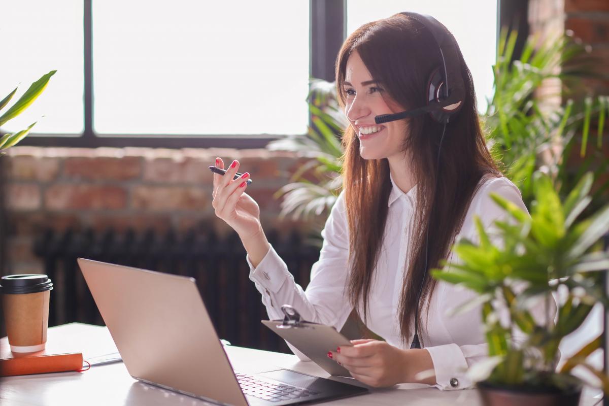 I migliori lavori da fare in smart working Segretaria ha in mano uno blocco note e una penna mentre parla al telefono usando un auricolare con microfono, seduta ad una scrivania con un computer portatile aperto